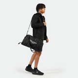 a fit man stand at right and wearing  tech zipped hoodie black with short black ,plus bag  black with logo, and shoes , 