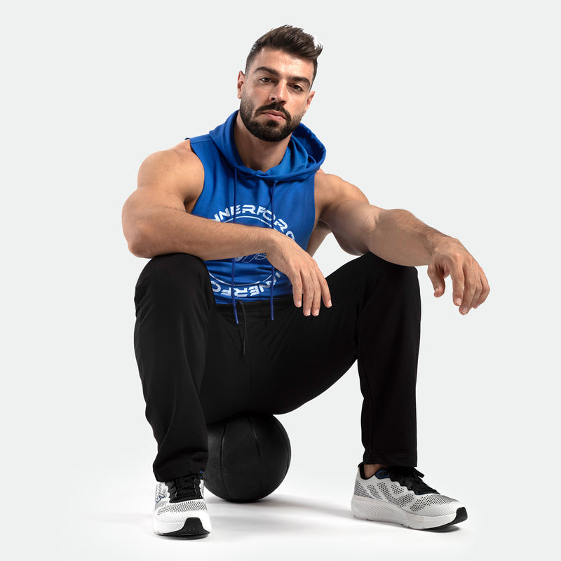 Man in a blue sleeveless hoodie and black pants sits confidently on a black exercise ball. White sneakers. Neutral background. Fitness theme.