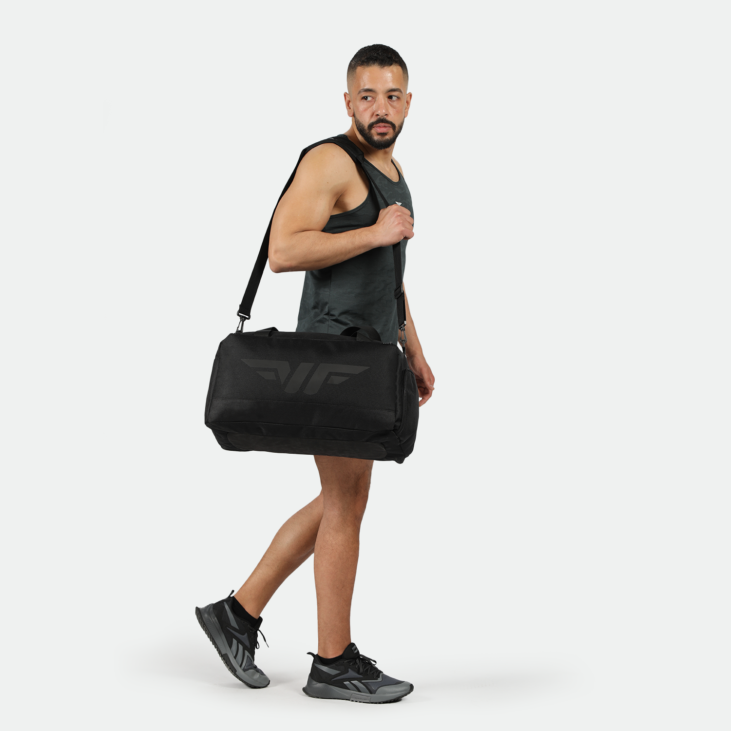 A man in a black tank top and shorts confidently walks, carrying a large black duffel bag over his shoulder. He wears black sneakers and is set against a plain white background.