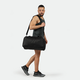 A man in a black tank top and shorts confidently walks, carrying a large black duffel bag over his shoulder. He wears black sneakers and is set against a plain white background.