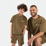 A smiling man and a child are wearing matching olive green outfits with "Winner Force" text. The man kneels while the child stands. The mood is playful.