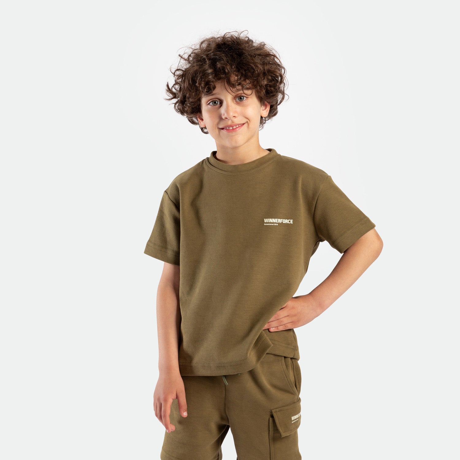 a young boy look at the camera and smile ,he wearing matching set olive drape , t-shirt with logo and short with tow pocket 