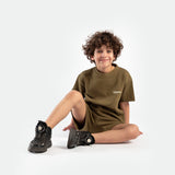 A smiling child with curly hair sits on the floor. Wearing an olive green outfit and black sneakers, the child exudes a relaxed, joyful vibe.