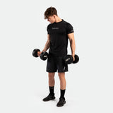 A man holding dumbbells wearing a performance fit t-shirt in the color black with the tri short, black socks, and black shoes.