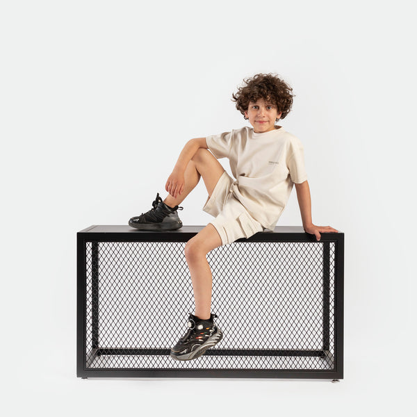 A young boy with curly hair sits confidently on a mesh table, wearing a beige outfit and black sneakers, against a white background.