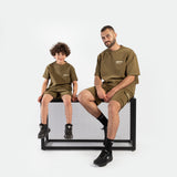 Father and son wearing matching olive green outfits, smiling warmly. The boy is standing, and the father is kneeling beside him, creating a joyful, familial tone.