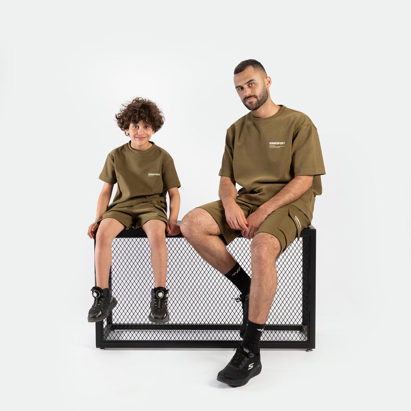Father and son wearing matching olive green outfits, smiling warmly. The boy is standing, and the father is kneeling beside him, creating a joyful, familial tone.