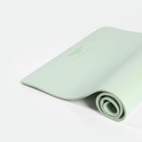 A light green yoga mat, partially rolled, lies on a white background. The mat features a minimalist logo, conveying a calm and serene vibe.