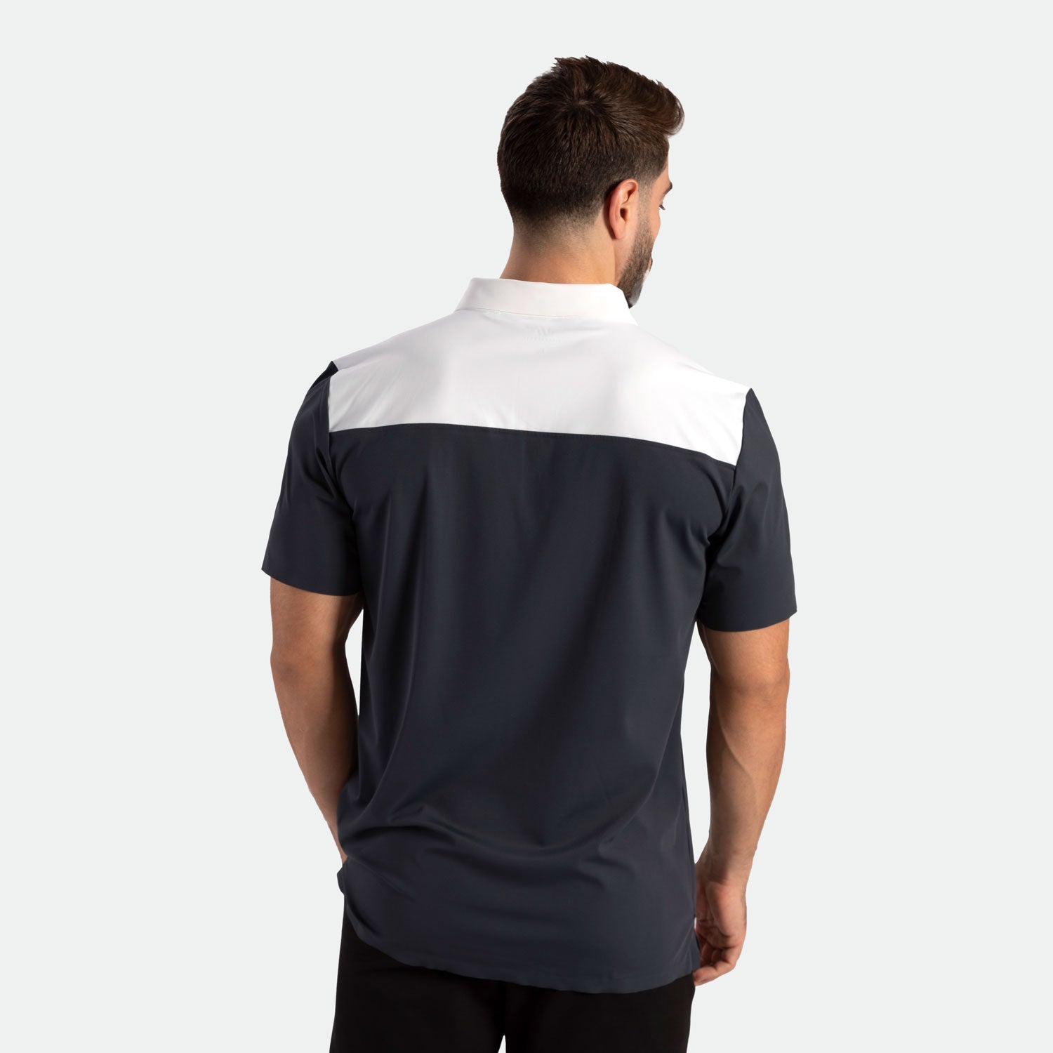 A man with short brown hair stands facing away, wearing a black and white short-sleeve polo shirt. The background is plain white, highlighting the clothing.