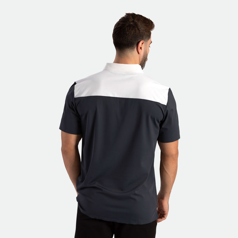 A man with short brown hair stands facing away, wearing a black and white short-sleeve polo shirt. The background is plain white, highlighting the clothing.