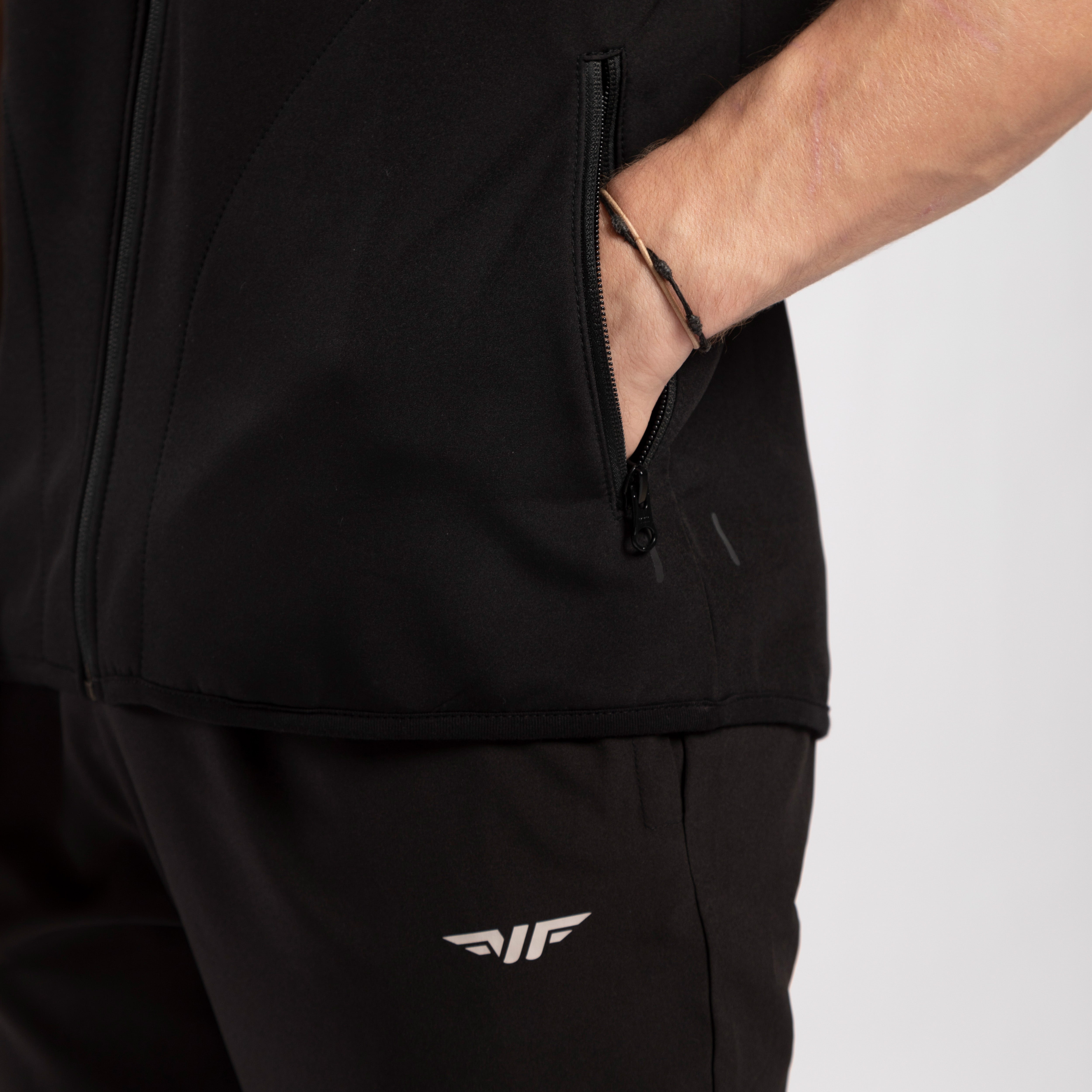 A person in black sportswear with a hand in the jacket pocket. The outfit features a white logo on the pants, conveying a sporty and casual tone.