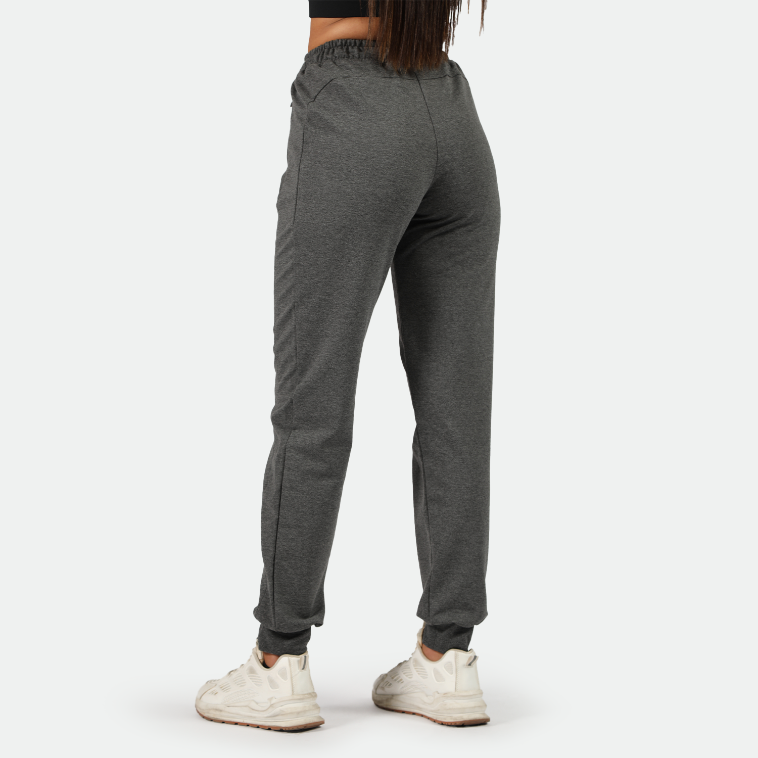 a back view for fit woman wearing dark grey  jogger pants with shoes white ,plus the background is light grey 