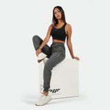 A woman wearing a black sports crop top and gray jogger pants with an elastic waistband, sitting on a white cube with the Runnerforce logo. She is wearing white athletic shoes and has straight, long hair.