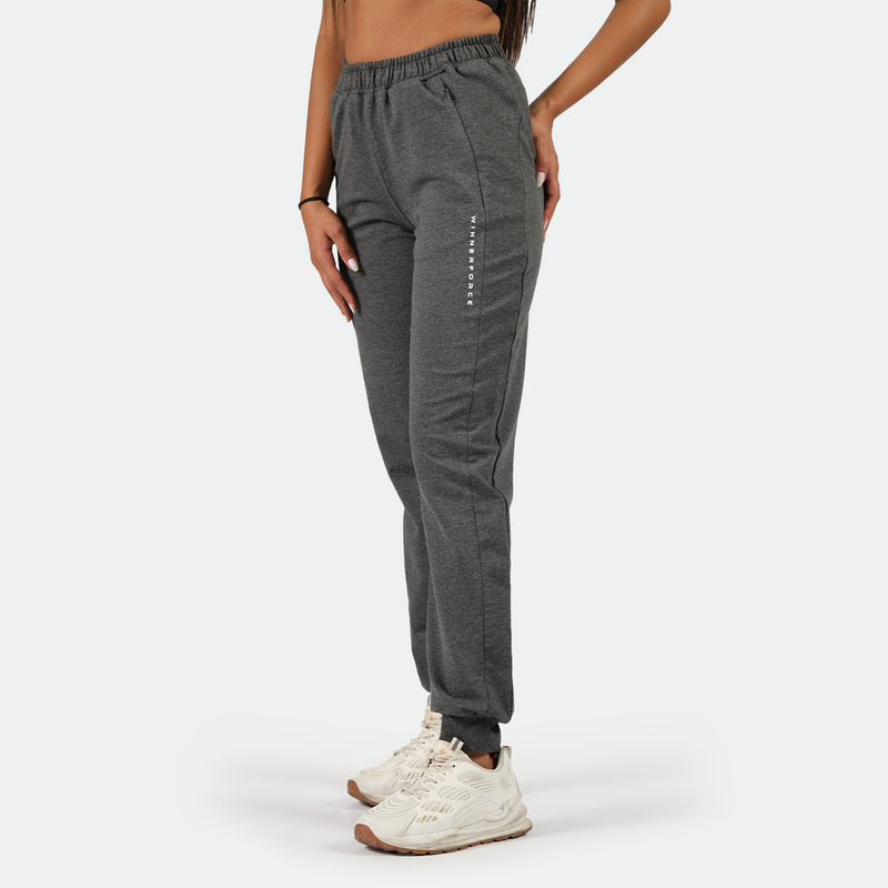 a fit woman stand at right and wearing dark grey jogger pants with logo ,she wearing shoes white ,plus the background is light grey 