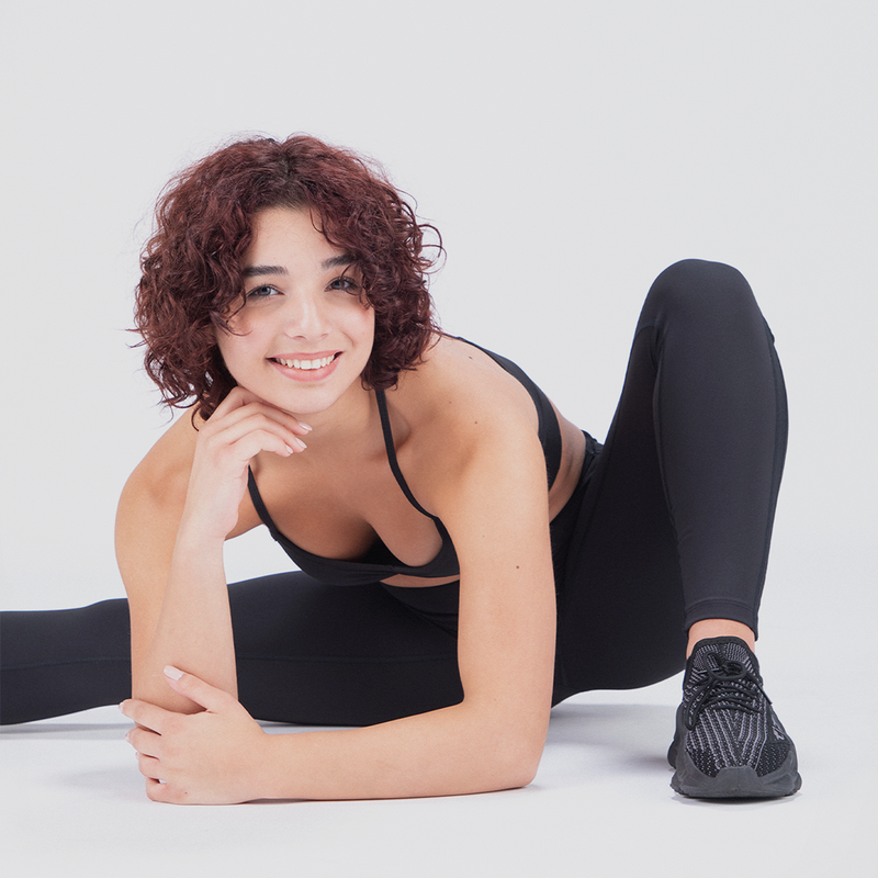 Woman with curly hair in black athletic wear smiles while stretching on the floor. Her relaxed pose conveys a cheerful and energetic mood.