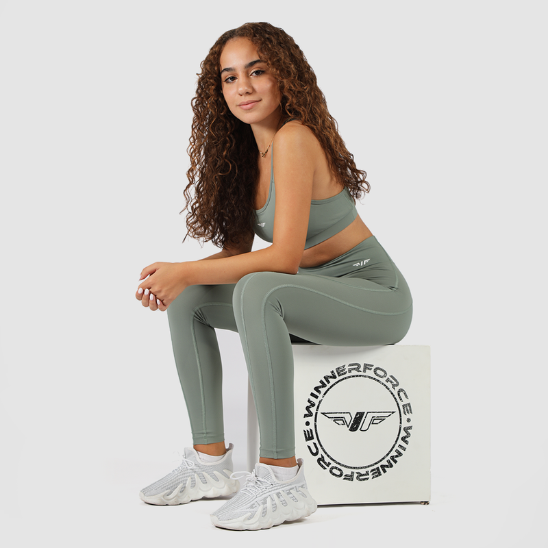 A young woman with curly hair is sitting on a white workout box, wearing an olive green sports bra and matching high-waisted leggings. She pairs the outfit with white athletic sneakers. She has a confident expression, looking directly at the camera with a relaxed posture. The background is plain white, and the box features a circular logo with wings and the text "WINNERFORCE