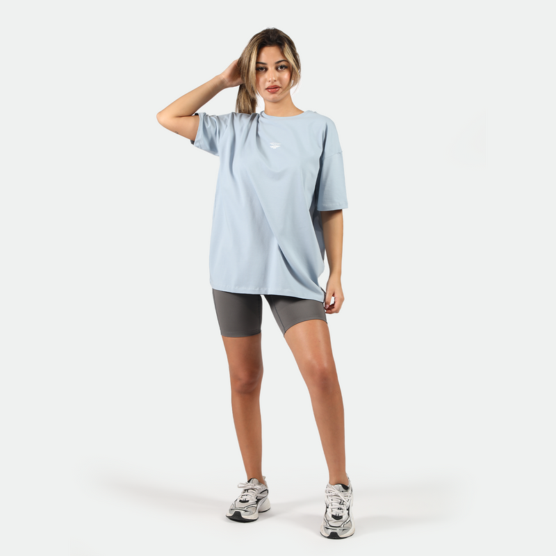 a fit woman stand with pose and look at the camera ,she wearing  t-shirt  blue with logo  and she wearing short grey and sneakers 