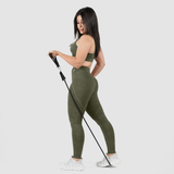 A woman with long, straight black hair is posing with a resistance band, wearing an olive green sports bra and matching high-waisted leggings. She pairs the outfit with white athletic sneakers. She stands in a three-quarter view, looking downward with a confident expression. The background is plain white.