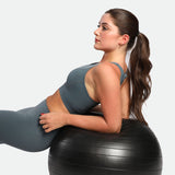 A woman in a gray sports outfit reclines on a black exercise ball. Her long hair is tied back, and she gazes upward, conveying focus and calm.