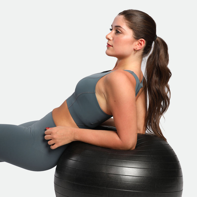 A woman in a gray sports outfit reclines on a black exercise ball. Her long hair is tied back, and she gazes upward, conveying focus and calm.