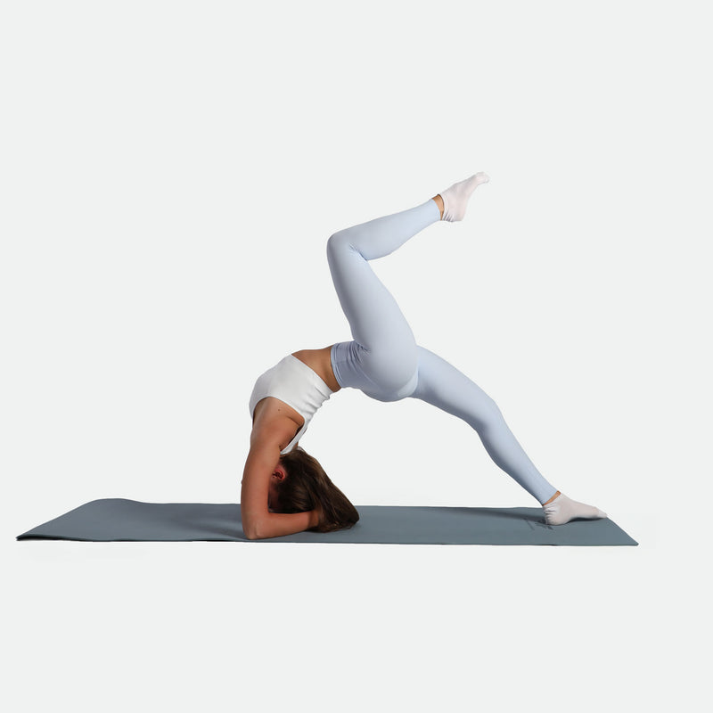 A person in a white top and leggings balances in a yoga pose on a mat, head down, one leg bent upward, conveying strength and focus.