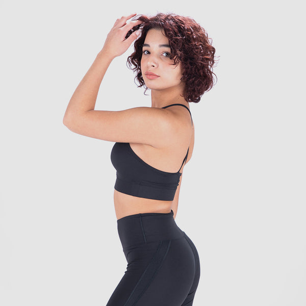 A person in a black sports bra and leggings poses sideways against a neutral grey background