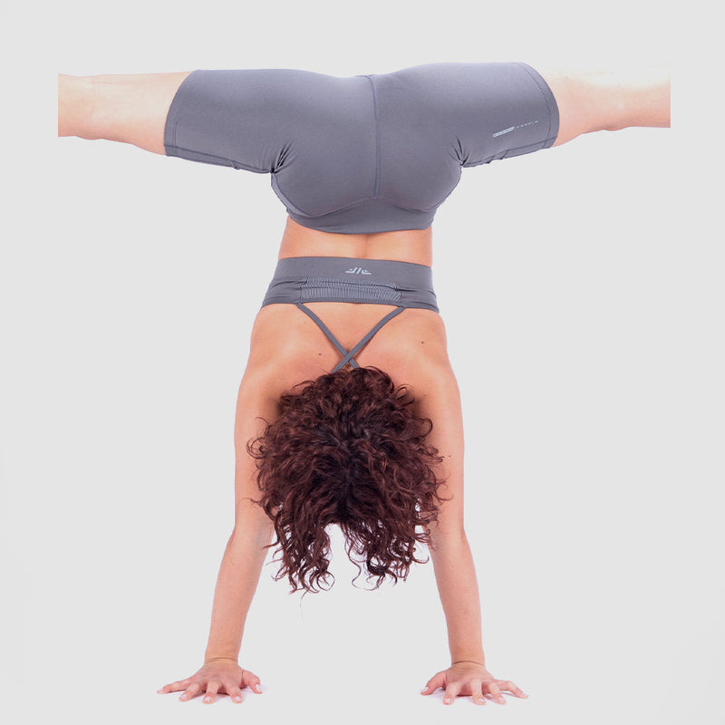 Person performing a handstand with legs spread wide apart, wearing a matching grey sports outfit consisting of a sports bra and fitted shorts