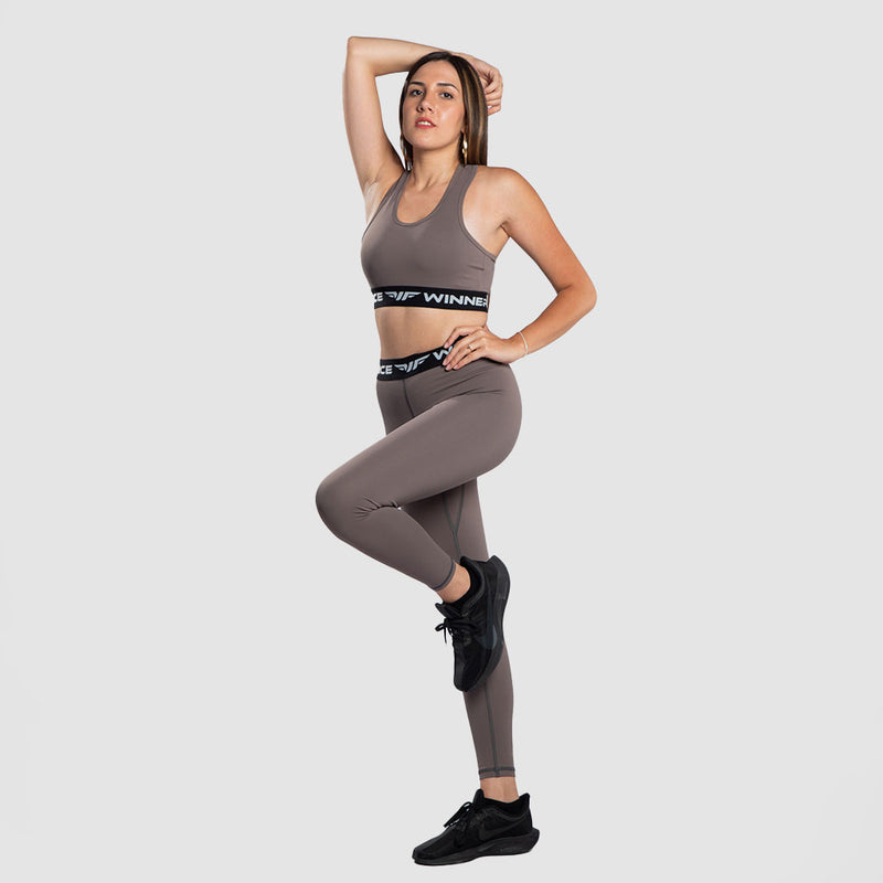 Woman posing in a taupe-colored athletic outfit with black sneakers, standing on one leg with one hand on her hip and the other resting on her head
