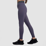 A woman with a fit physique stand at right  ,she wearing a purple  legging  with black elastic bands featuring white text, She is also wearing black athletic shoes ,plus the background is light grey 