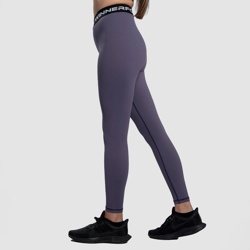 A woman with a fit physique stand at right  ,she wearing a purple  legging  with black elastic bands featuring white text, She is also wearing black athletic shoes ,plus the background is light grey 