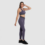 A woman with a fit physique is wearing a matching purple sports bra and leggings set with black elastic bands featuring white text. She is posing in a side profile with one hand touching her hair. She is also wearing black athletic shoes. The background is  light grey