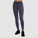 A woman with a fit physique is wearing a matching purple sports bra and leggings set with black elastic bands featuring white text. She is posing in a side profile with one hand touching her hair. She is also wearing black athletic shoes. The background is  light grey