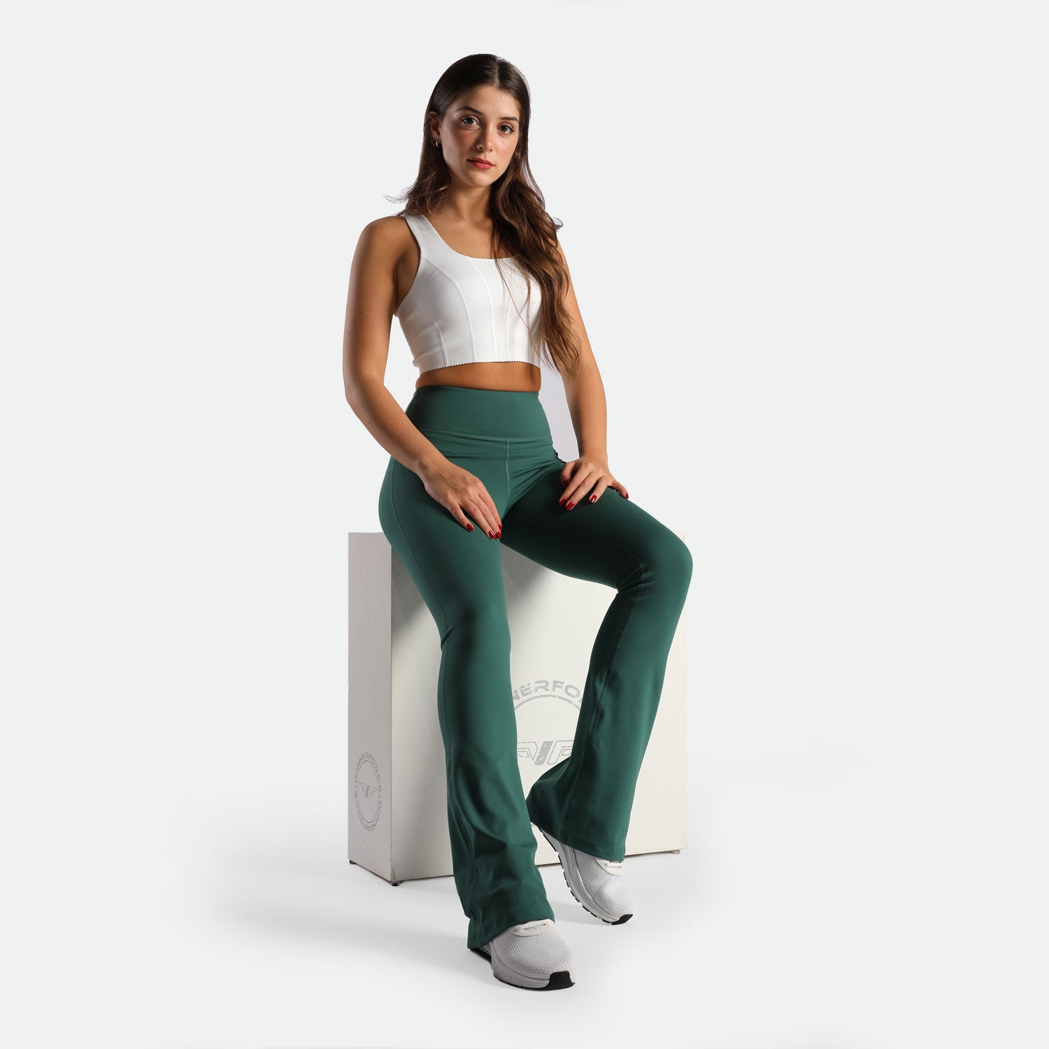 Young woman in a white crop top and green leggings sits on a white cube. She appears confident and relaxed against a plain background.