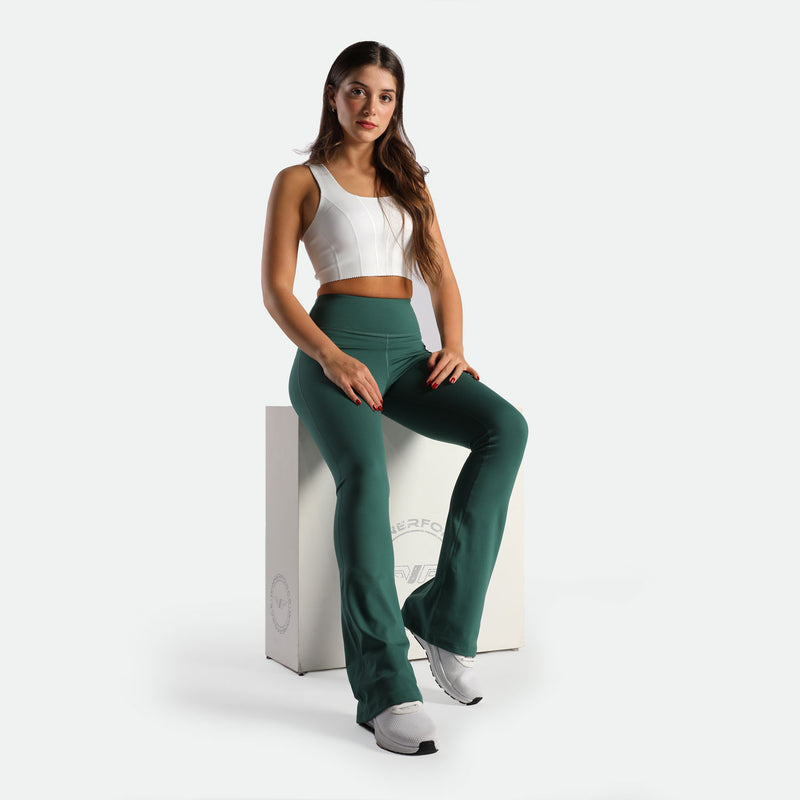 Young woman in a white crop top and green leggings sits on a white cube. She appears confident and relaxed against a plain background.