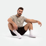 Seated man wearing a beige t-shirt, black shorts, and white socks on a light background. He appears relaxed and focused, conveying a calm mood.
