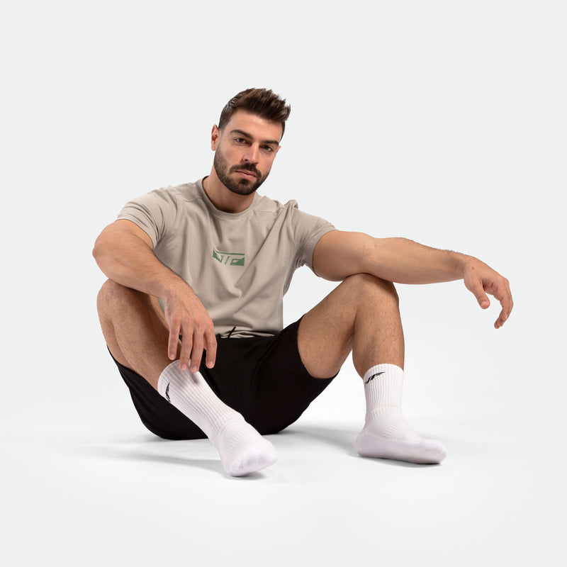Seated man wearing a beige t-shirt, black shorts, and white socks on a light background. He appears relaxed and focused, conveying a calm mood.