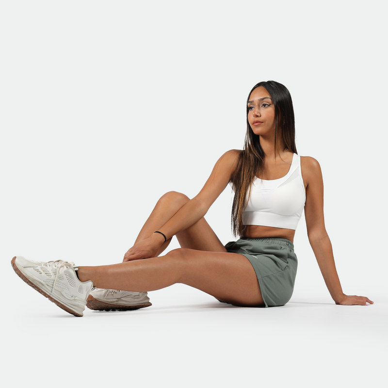 A woman wearing a white sports bra and green athletic shorts, sitting on the floor with one leg bent and the other extended. She is wearing white sneakers and has long, straight hair. The background is plain white.
