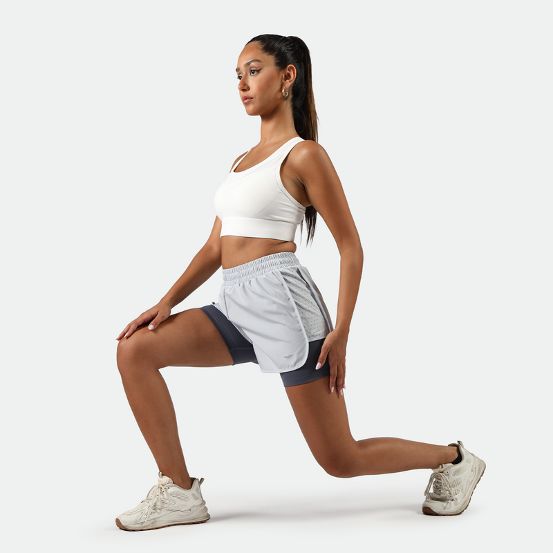 A woman wearing a white sports bra and light gray athletic shorts with a darker inner layer, performing a lunge exercise. She is wearing white sneakers and has her hair tied back in a ponytail. The background is plain white.