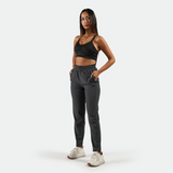 a fit woman  look at the camera and wearing bra black and pants grey with  white  athletic shoes ,plus the background is light grey 