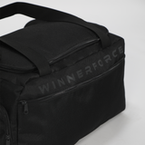 Black duffel bag against a light background. It features zippers, short handles, and a shoulder strap, conveying a sleek and functional design.