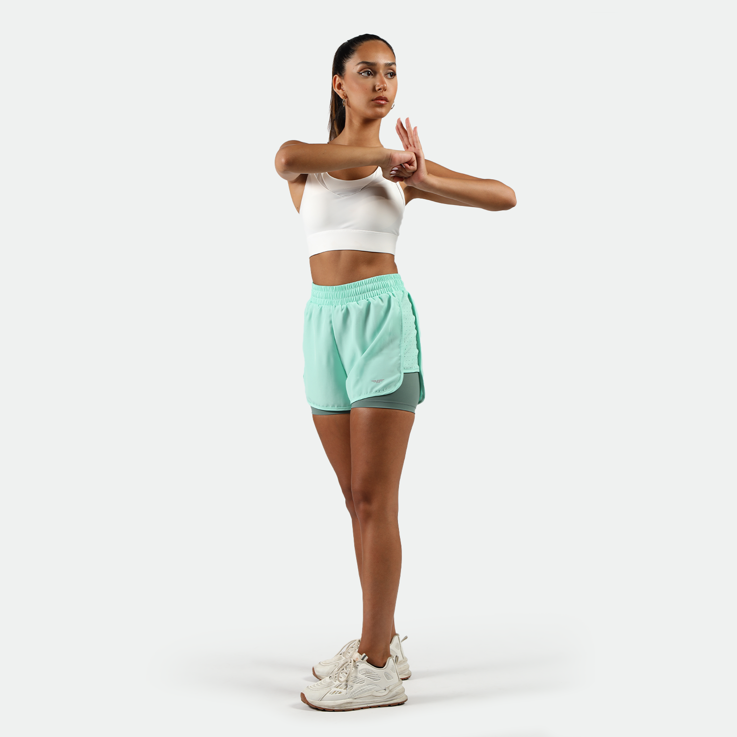 a fit woman stand at right  with pose ,and she wearing sports bra white and  beach glass blue with shoes white ,plus the background is light grey 