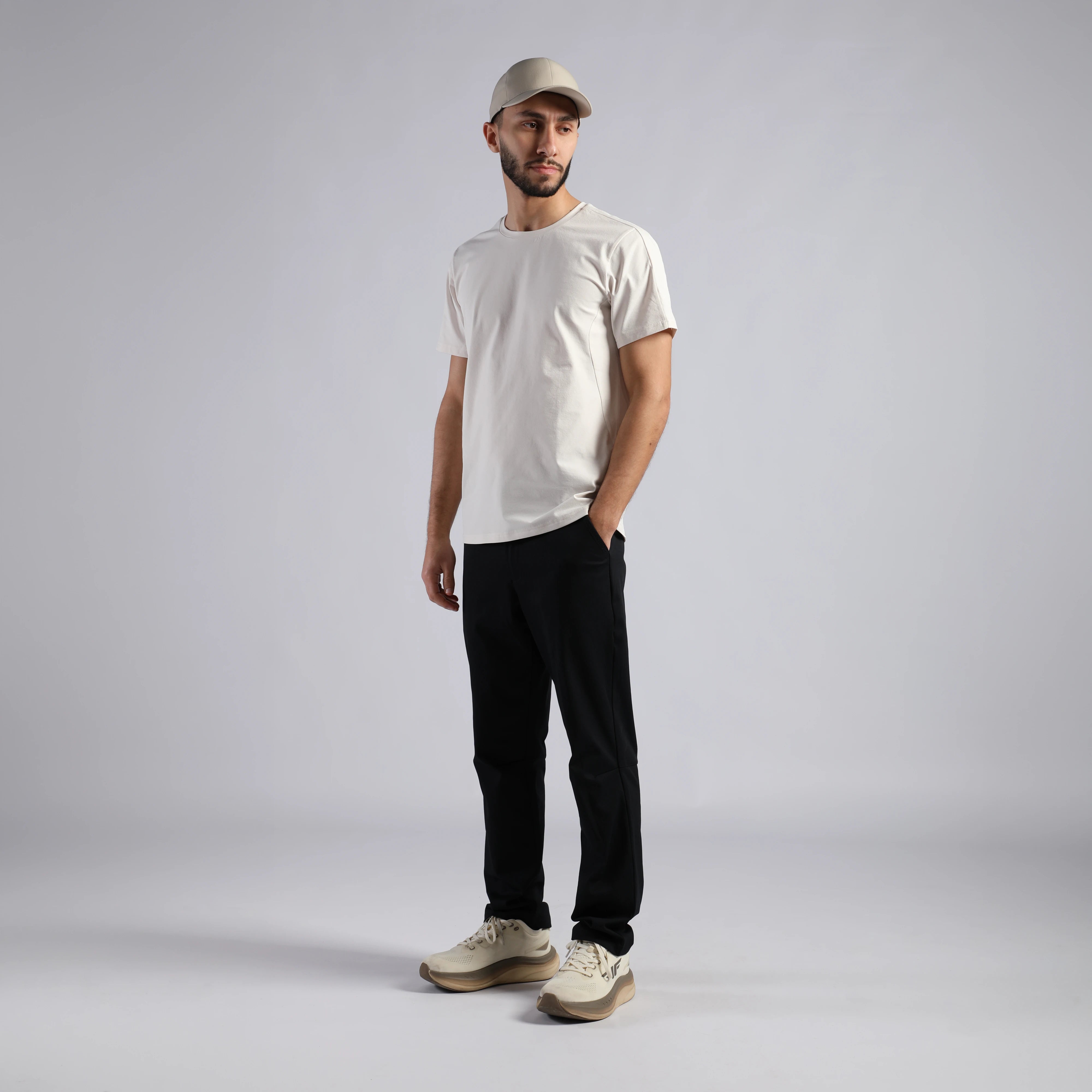 A man modeling a white crew-neck t-shirt, black pants, beige sneakers, and a beige baseball cap against a light gray studio background.