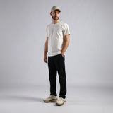 A man modeling a white crew-neck t-shirt, black pants, beige sneakers, and a beige baseball cap against a light gray studio background.