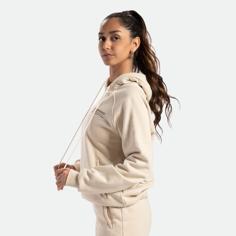 a fit woman stand at right and wearing  hoodie  and pants beige 