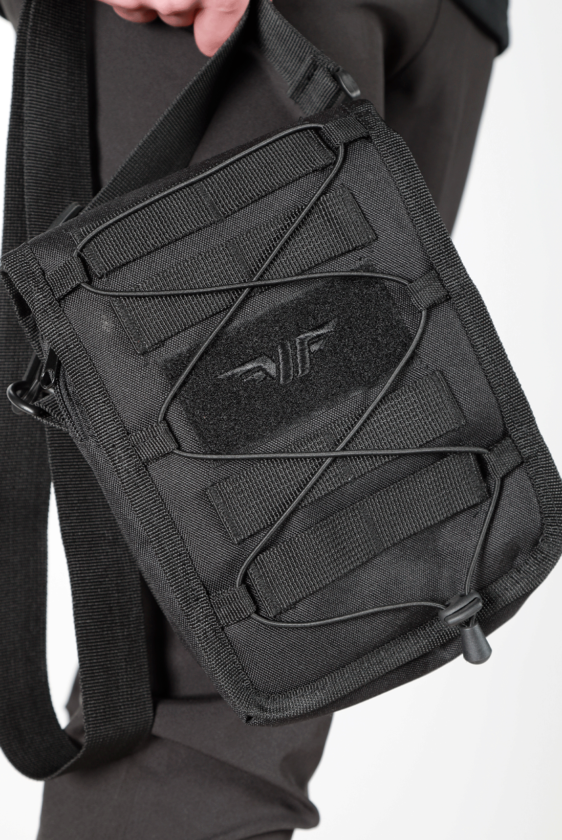 close up view  for  small, black crossbody shoulder bag with an adjustable strap, front elastic cord, and a subtle logo on the front, displayed against a plain light gray background.