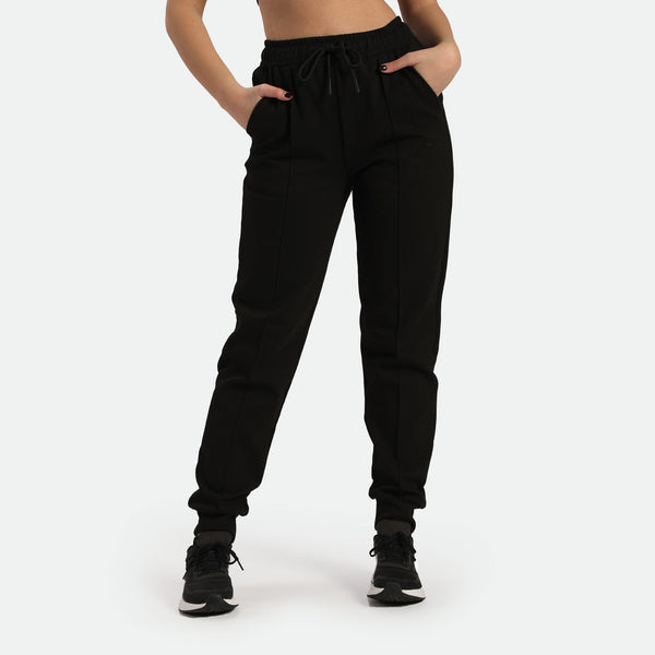 a fit woman stand with pose, she wearing black jogger pants with shoes ,plus the background is light grey 