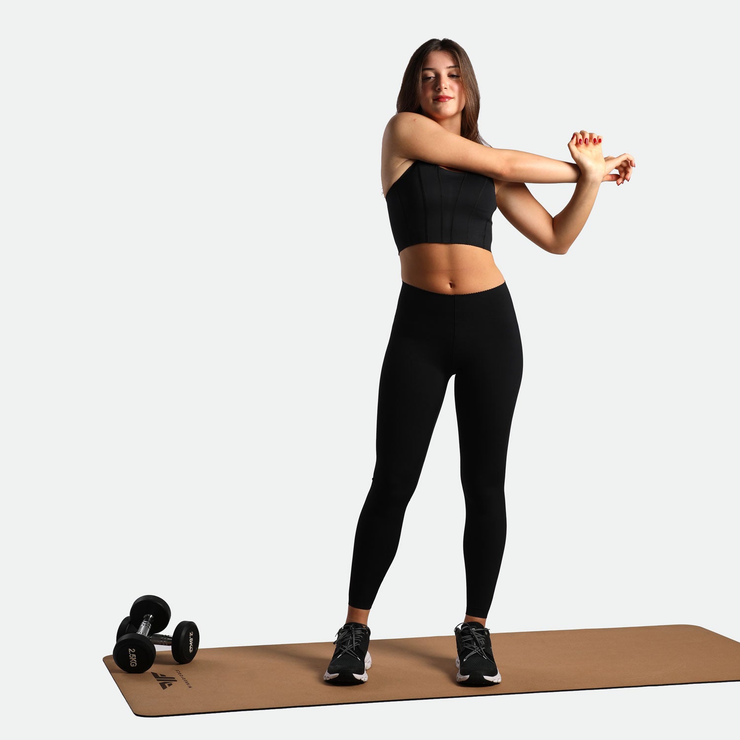 A woman in black workout attire stretches her arms on a yoga mat, with dumbbells beside her, conveying a sense of fitness and determination.