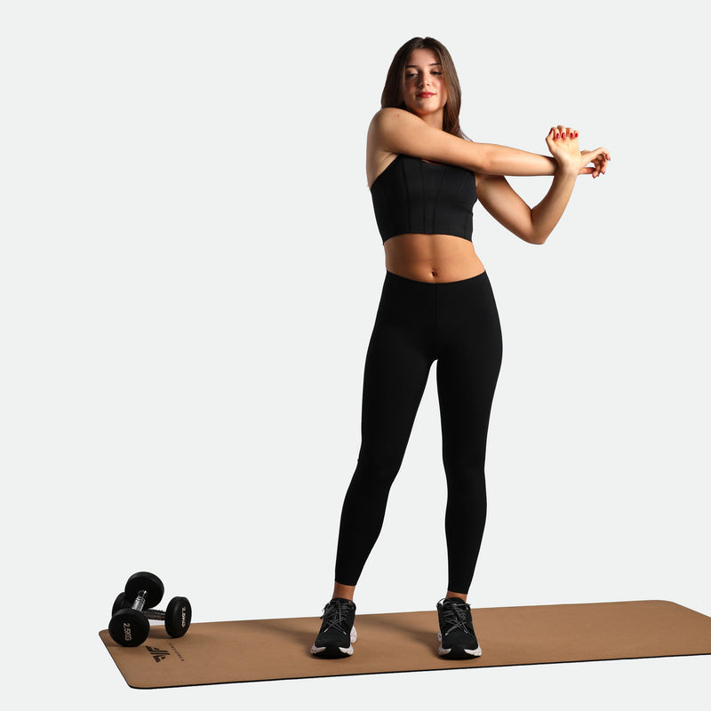 A woman in black workout attire stretches her arms on a yoga mat, with dumbbells beside her, conveying a sense of fitness and determination.