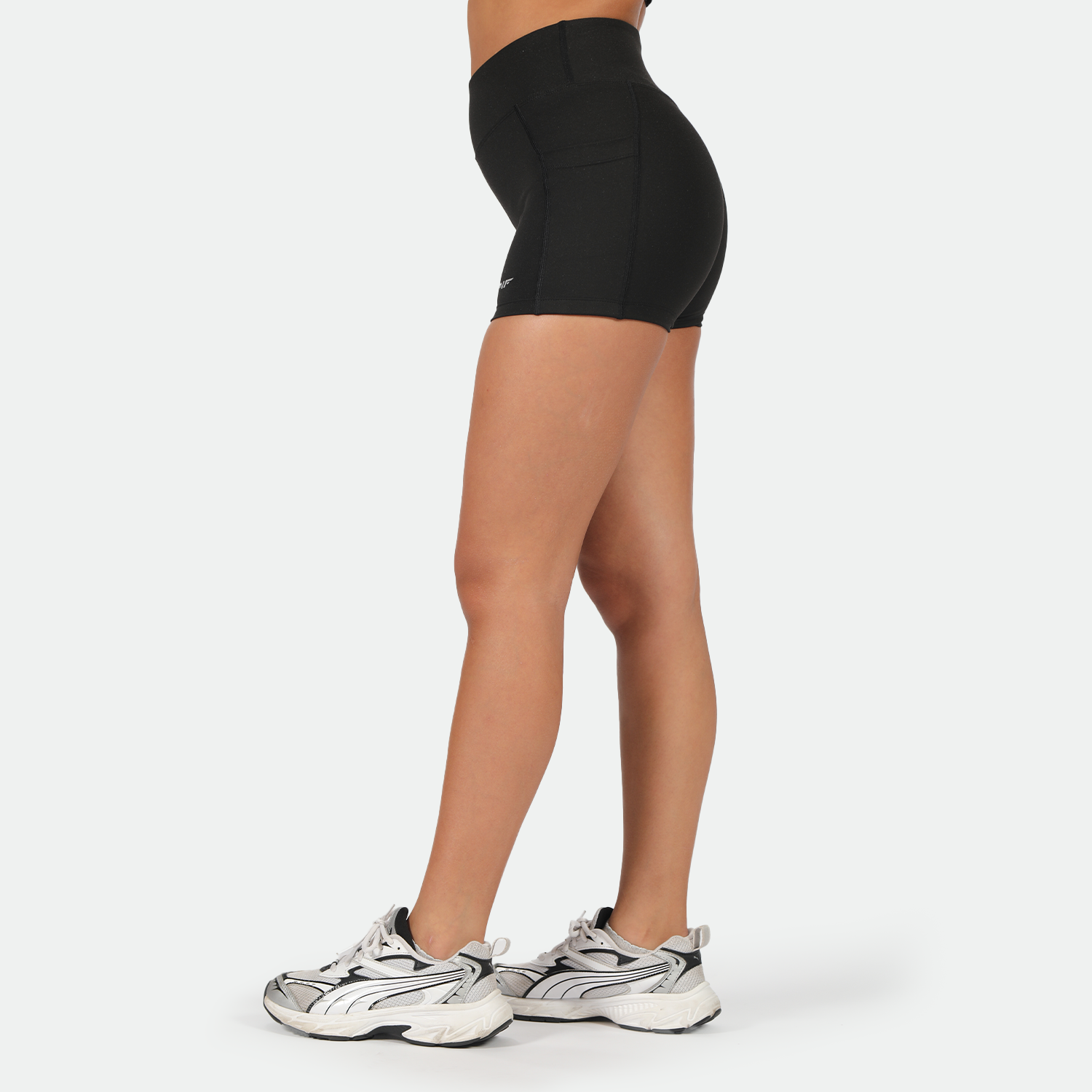 a fit woman stand at right wearing short black with shoes ,plus the background is light grey 