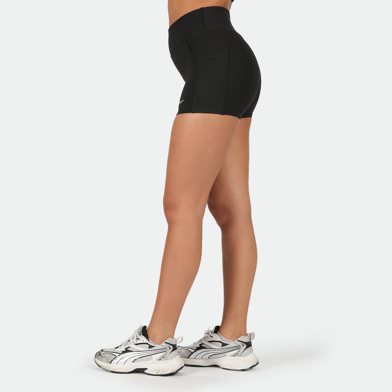 a fit woman stand at right wearing short black with shoes ,plus the background is light grey 